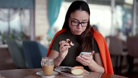 Cute Adult Woman Wearing Glasses Eating and Browsing a Mobile App alt