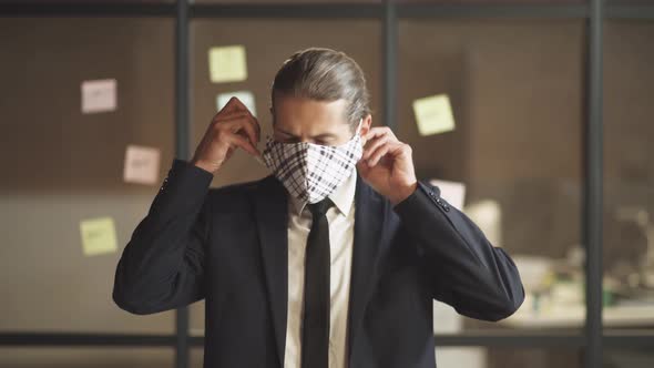 Business Meeting, Young Man in Suit Stands in Conference Room, Man Puts on a Cloth Mask To Protect alt