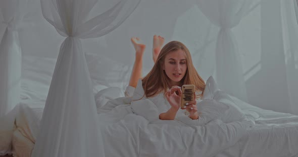 Nice Girl Lies on a Bed in a Cozy Bedroom and Communicates By Video Call with Her Friend alt