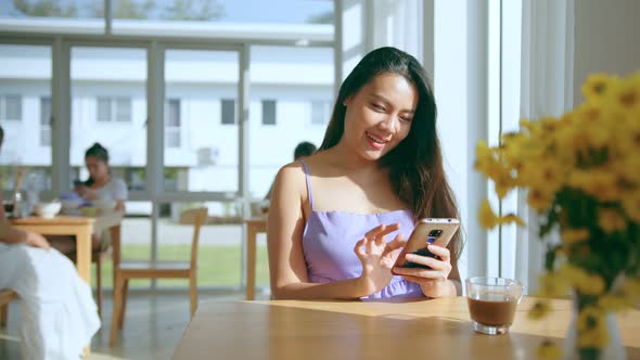 young attractive asian female hand hold smartphone sit relax at dining table near window