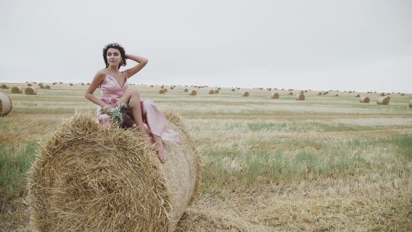 Elegant Lady in Dress Posing with Smile for Camera on a Haystack in Windy Field alt