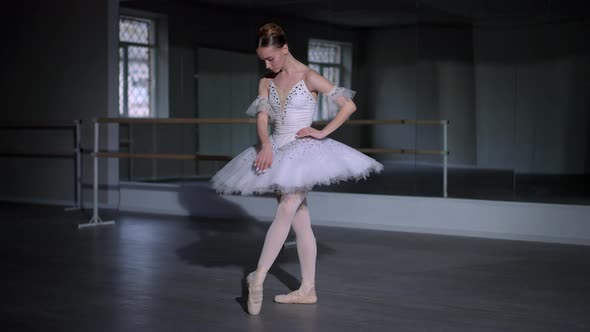 Graceful Confident Ballerina in Fourth Position Standing in Dark Dance Studio Adjusting White Tutu alt