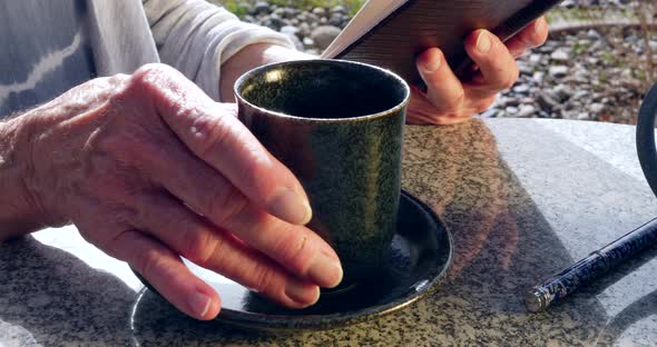 Close up on an elderly woman reading a book and drinking a cup of herbal tea in the morning sun. alt