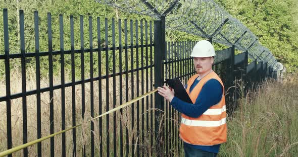 Worker with a Measuring Tape Measure a Fence with Barbed Wire alt