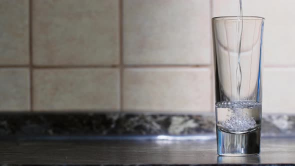 A transparent liquid is poured into a glass on a table against the backdrop of a kitchen wall. Vodka