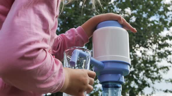 a Little Girl in a Pink Jacket with Difficulty Pumps Water Into a Glass By Pressing the Pump on a