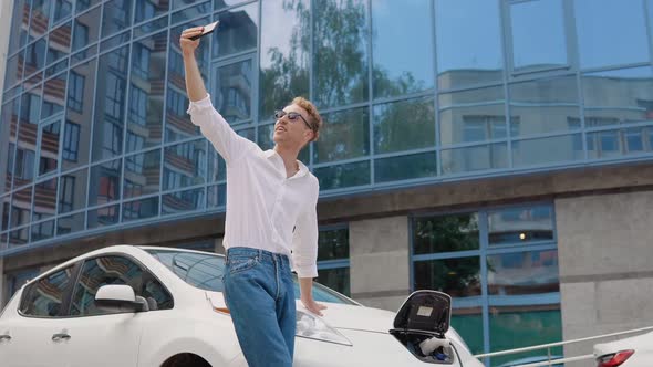 Stylish Modern Young Curly Man Stands Near an Electric Car Charging and Takes a Selfie with His alt