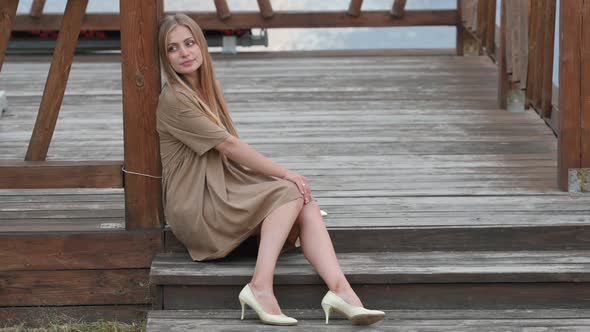 Young attractive blonde girl in a dress sits on a wooden pier
