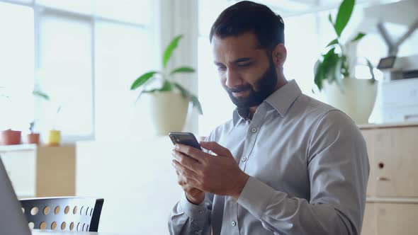Happy Indian Business Man Using App on Cell Phone Working in Office alt