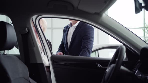Professional Car Dealer Opening Vehicle Doors for Wealthy Senior Caucasian Woman in Showroom. Rich alt