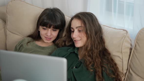 Couple of Beautiful Cheerful Young Women Smiling While Watching Film on Computer on Sofa alt