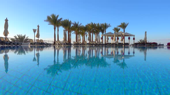 Swimming pool with clear blue water, palm trees, deck chairs, gazebo and clean blue sky.