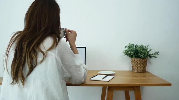 young smiling woman working on laptop while enjoying drinking warm coffee sitting in a living room. alt