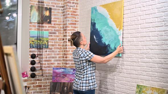 A girl hangs a picture on the wall in her creative studio, then moves away alt