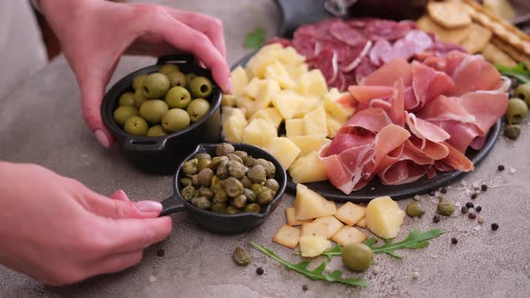 Making Meat and Cheese Antipasto Plater Woman Adding Capers and Olives to Serving Board with Cheese alt