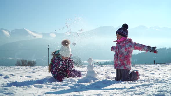 Children in Outwear Making Small Snowman While Playing on Snowy Field in Sunlight with Mountains on