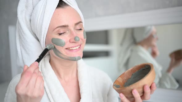 Beautiful Smiling Woman at Home in Bathrobe with a Towel Applying Face Clay Mask Against Acne To
