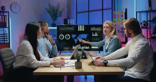 Teammates Browsing World Statistic Indicators on TV Screen During Joint Meeting in Evening Office alt