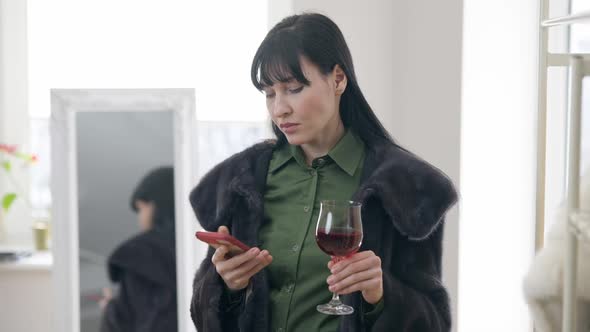 Portrait of Narcissistic Young Woman in Fur Coat Holding Glass with Wine Calling on Smartphone alt