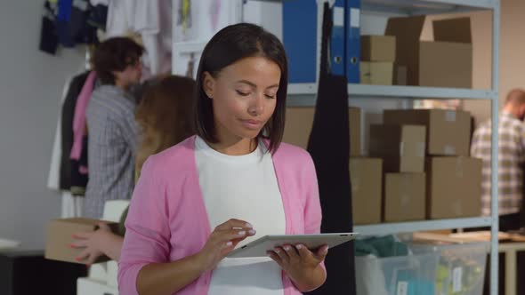 Afroamerican Woman Retail Seller with Digital Tablet Smiling at Camera in Clothes Warehouse alt
