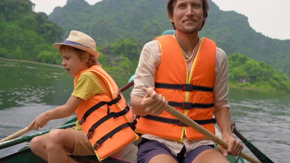 A Young Man and His Son on a Boat Rowing Together on a River Trip Among Spectacular Limestone Rocks alt
