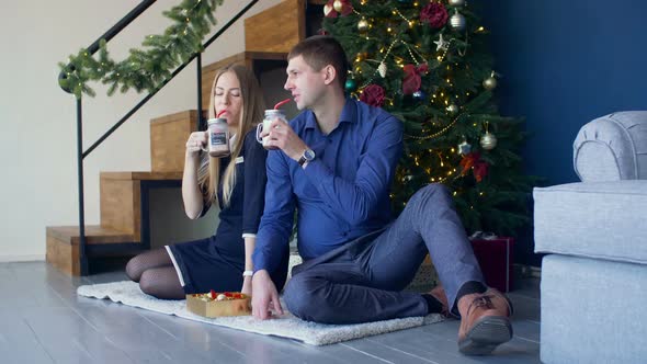 Happy Couple Enjoying Hot Drink Under Christmas Tree alt
