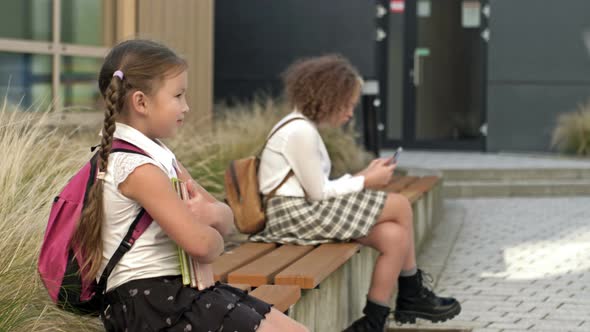 Two Schoolgirls of Different Ages are Sitting on a Bench in the School Yard