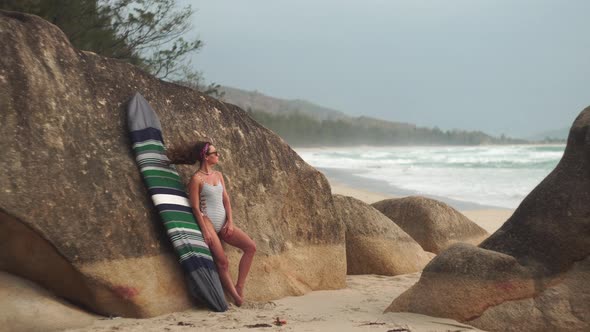 Woman Poses with Surfboard in Case Leaning Against Rock