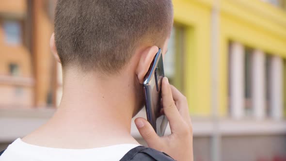 A Caucasian Teenage Boy Talks on a Smartphone  Rear Closeup  a School in the Background