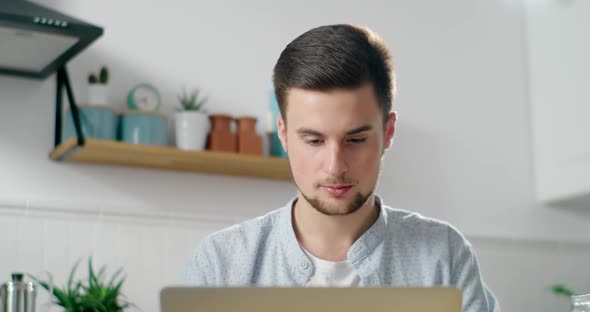 Young Bearded Man Works Home on Laptop in Kitchen, Drinking Coffee in Morning