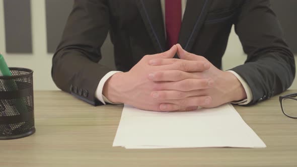 Unrecognizable Man in the Suit Sitting at the Table with Crossed Fingers in Front of Blank Papers alt