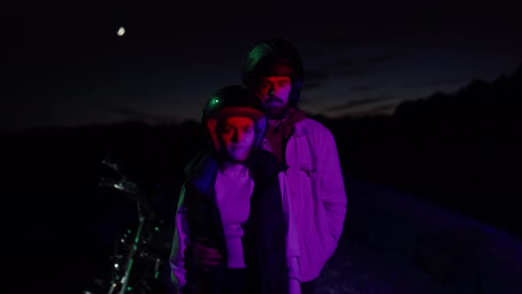 Couple Looking At Camera, Wearing Helmet And Standing Next To A Motorbike At Night. - medium shot alt
