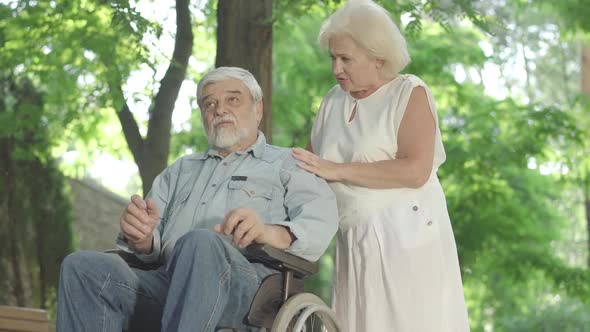Bottom Angle View of Handicapped Old Man and Caring Senior Woman Talking in Sunlight in Summer Park alt