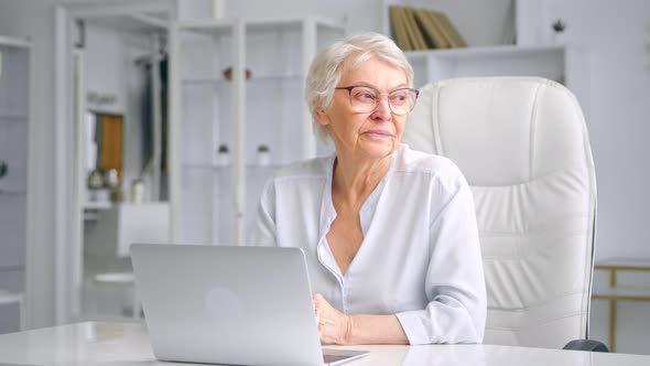 Senior woman with short grey hair and glasses glances at laptop display alt