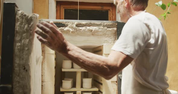 Senior caucasian man wearing apron and firing pottery in kiln at pottery workshop alt