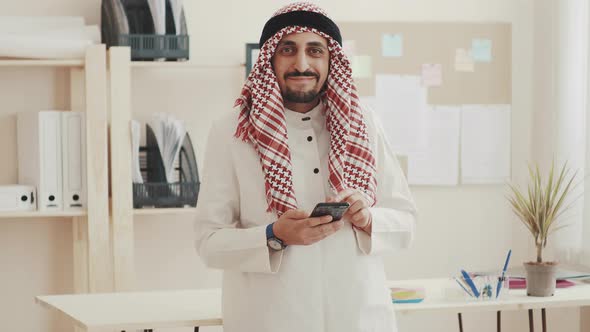 The Sheikh in Kufiyeh Looks at Phone, Looks at the Camera, and Nods. The Arab Businessman at the alt