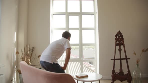 A man in spacious room gets up from the sofa and goes to think to the big window alt