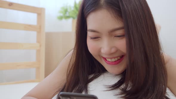Young Asian woman using smartphone checking social media while lying on bed in the morning. alt