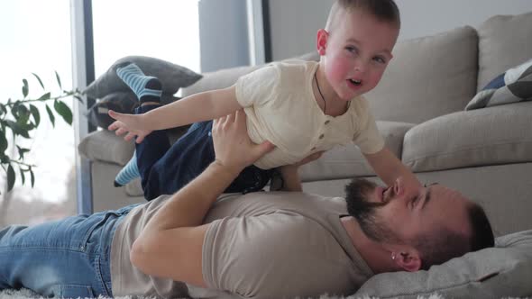 Loving dad lifting cute small kid son play plane having fun on sofa in living room, happy family adu