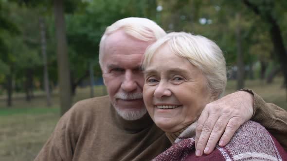 Senior Husband Hugging His Mature Wife Sitting on Bench in Park Posing to Camera