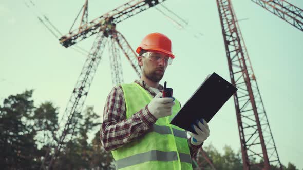 Electrical Equipment Worker Near High Voltage Tower Using Walkie Talkie and Clipboard alt