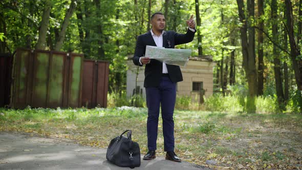 Wide Shot of Positive African American Man in Suit Standing in Summer Suburban Park with Paper Map alt