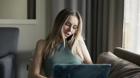 Blonde Woman Sitting on Sofa with Laptop on Knees and Talking By Smartphone alt