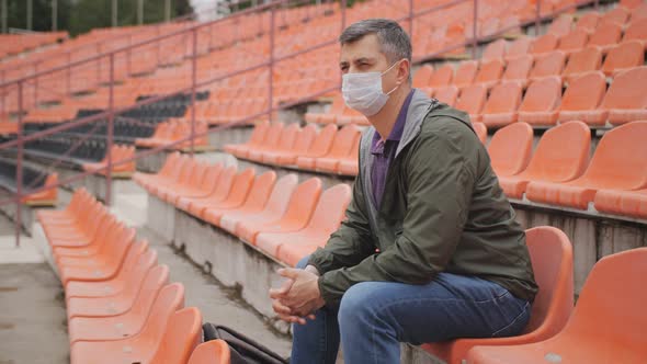 A Sad Middleaged Man Sits in the Stands of an Empty Stadium Wearing a Protective Medical Mask