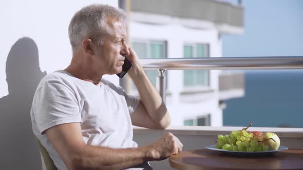 An Elderly Whitehaired Man is Talking on the Phone on the Balcony Eating Fruit and Enjoying the Sea