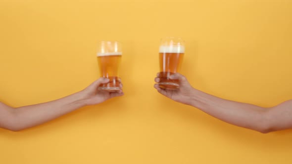 Hands of two friends holding and clanging glasses of beer on isolated yellow background