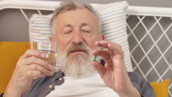 Caucasian Senior Man Taking Medicines and Drinking Water While Lie on Bed Alone