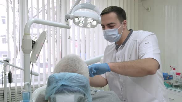 Young Male Dentist in Protective Mask Working with Elderly Patient in Dental Clinic alt