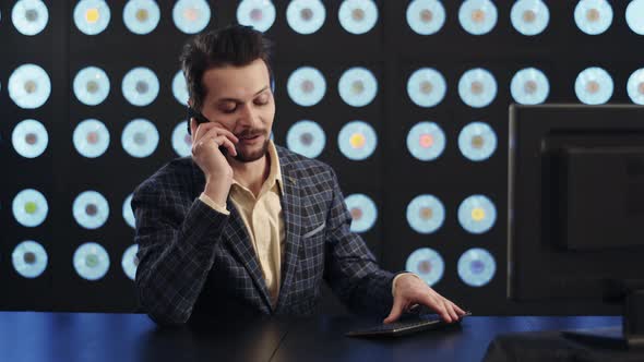 Stylish Businessman in Checkered Suit Sits at PC Desktop Speaking on Smartphone