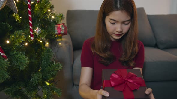 Young Asia female having fun opening X'Mas present box near Christmas tree decorated with ornaments. alt